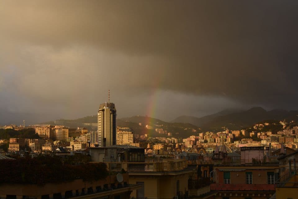 Un arcobaleno si estende su una città soleggiata tra le colline, sotto un cielo di nuvole scure e pesanti.