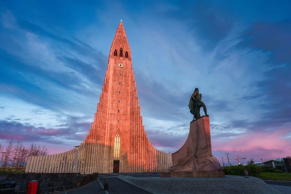 Eine hohe, modernistische Kirche und eine Statue auf einem Sockel, beleuchtet vom warmen Licht des Sonnenuntergangs vor einem bewölkten blauen Himmel.