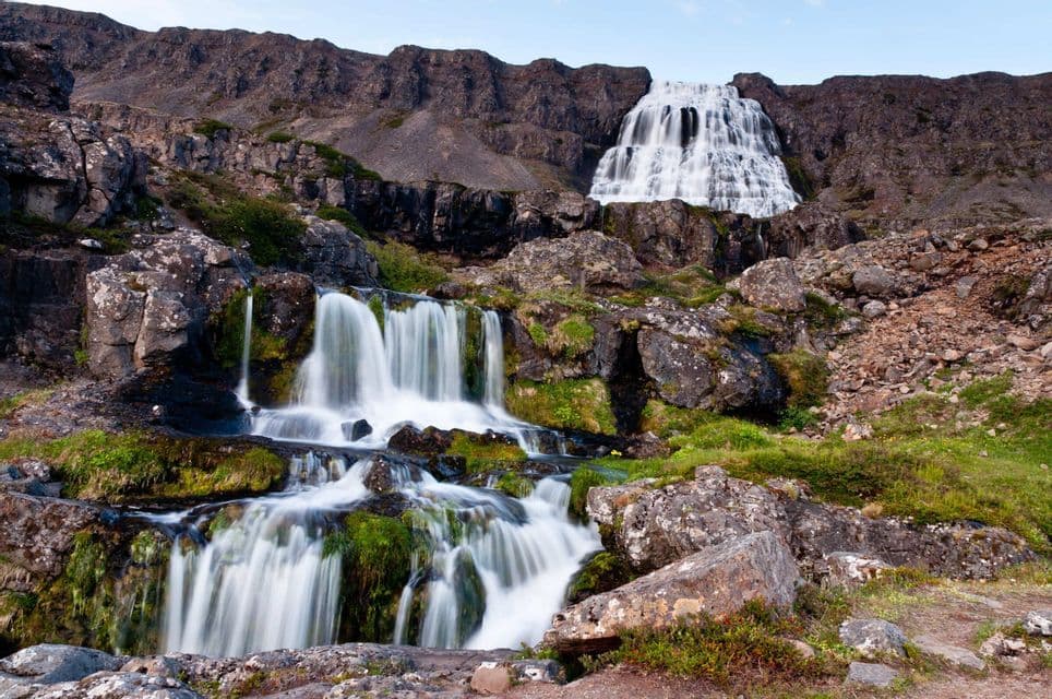 Eine Langzeitbelichtungsaufnahme von zwei Wasserfällen an einem felsigen, moosbewachsenen Berghang unter klarem Himmel.