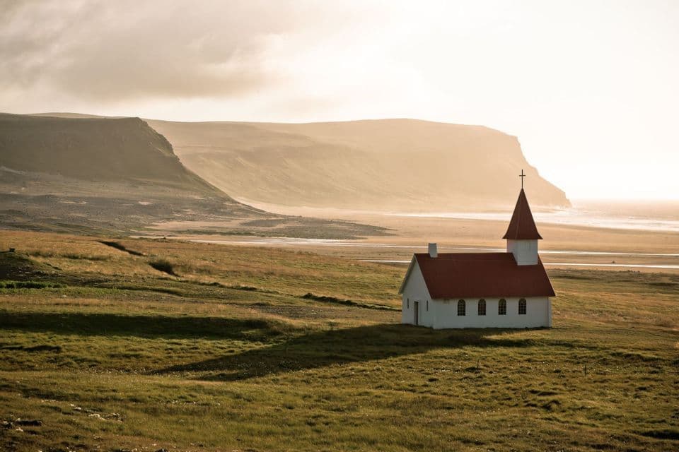 Eine einsame weiße Kirche mit rotem Dach steht auf einem grünen Feld, mit verschwommenen Klippen und einer Küstenlinie in der Ferne.