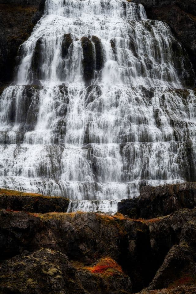 Nahaufnahme eines großen, mehrstufigen Wasserfalls, der über dunkle Felsen stürzt, mit herbstlich gefärbter Vegetation an seiner Basis.