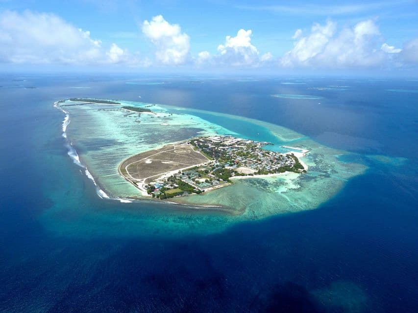 Una vista aérea de una isla tropical poblada rodeada de aguas de arrecife turquesas y el mar azul profundo bajo un cielo parcialmente nublado.