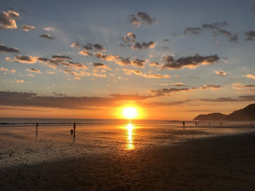 Un tramonto dorato si riflette sulla sabbia bagnata di una spiaggia, con le silhouette delle persone che guardano dalla riva sotto un cielo parzialmente nuvoloso.