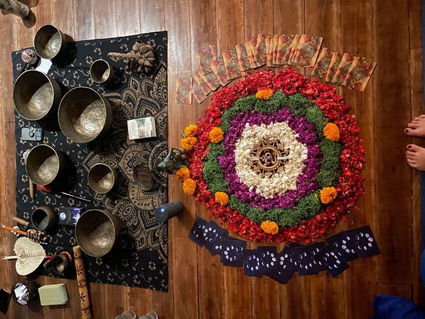 Una vista dall'alto di campane tibetane, un mandala floreale colorato e carte oracolo disposte su un pavimento di legno per una cerimonia spirituale.