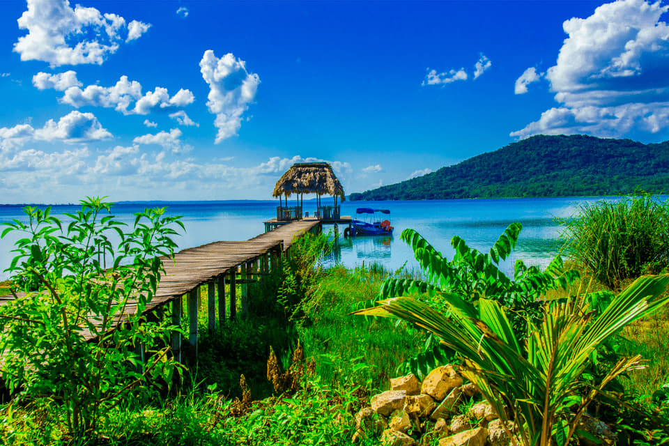 Ein Holzsteg führt zu einer reetgedeckten Hütte über einem ruhigen blauen See, mit einem bewaldeten Hügel und einem strahlend blauen Himmel im Hintergrund.