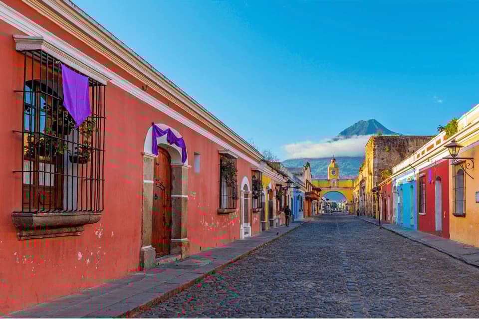 Eine Kopfsteinpflasterstraße mit bunten Gebäuden führt zu einem gelben Torbogen, im Hintergrund ist ein großer Vulkan sichtbar.