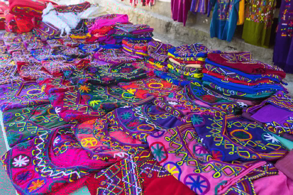 Nahaufnahme eines Marktstandes mit leuchtend bunten, aufwendig bestickten Textilien und Kleidungsstücken, die in Stapeln liegen.
