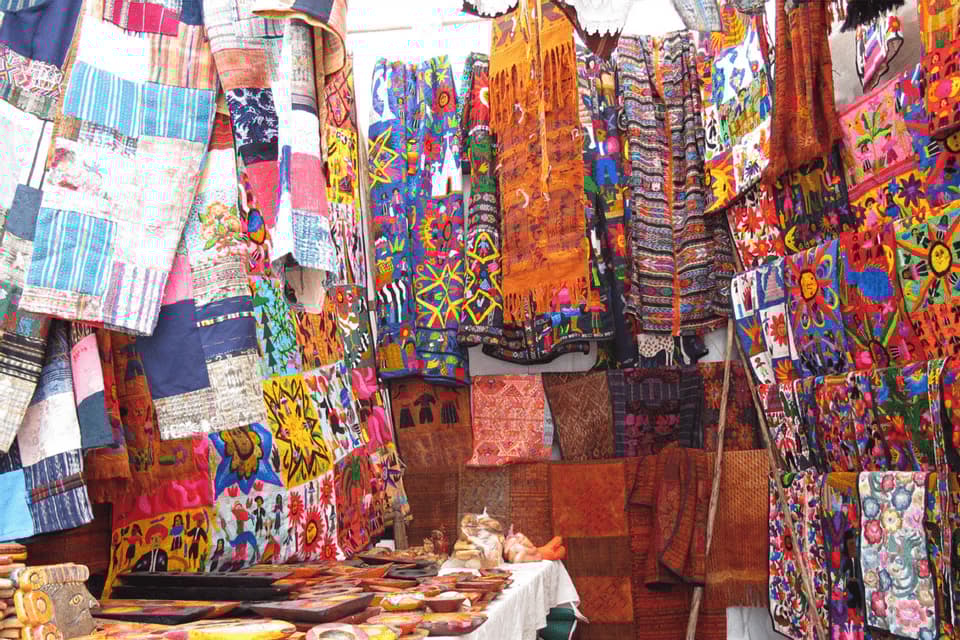 Eine Vielzahl farbenfroher, gemusterter Textilien und Kunsthandwerke wird an einem Marktstand im Freien zum Verkauf angeboten.