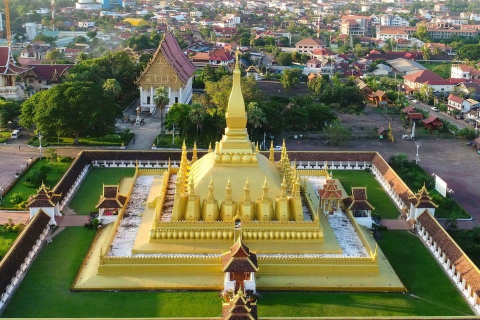 Eine Luftaufnahme einer großen goldenen Stupa in einem ummauerten Hof mit grünen Rasenflächen, im Hintergrund erstreckt sich eine Stadtlandschaft.