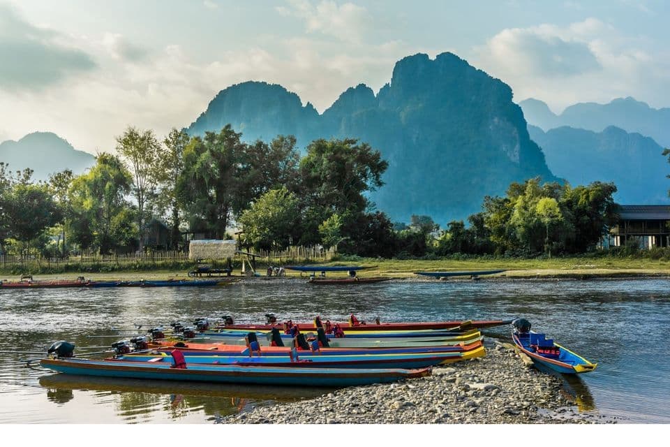 Bunte Langschwanzboote liegen an einem felsigen Flussufer vor Anker, mit üppig grünen Hügeln und großen Bergen in der Ferne.