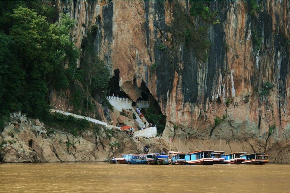 Langschwanzboote liegen an einem braunen Fluss am Fuße einer großen Klippe, wo Menschen weiße Treppen zu einem Höhleneingang hinaufsteigen.
