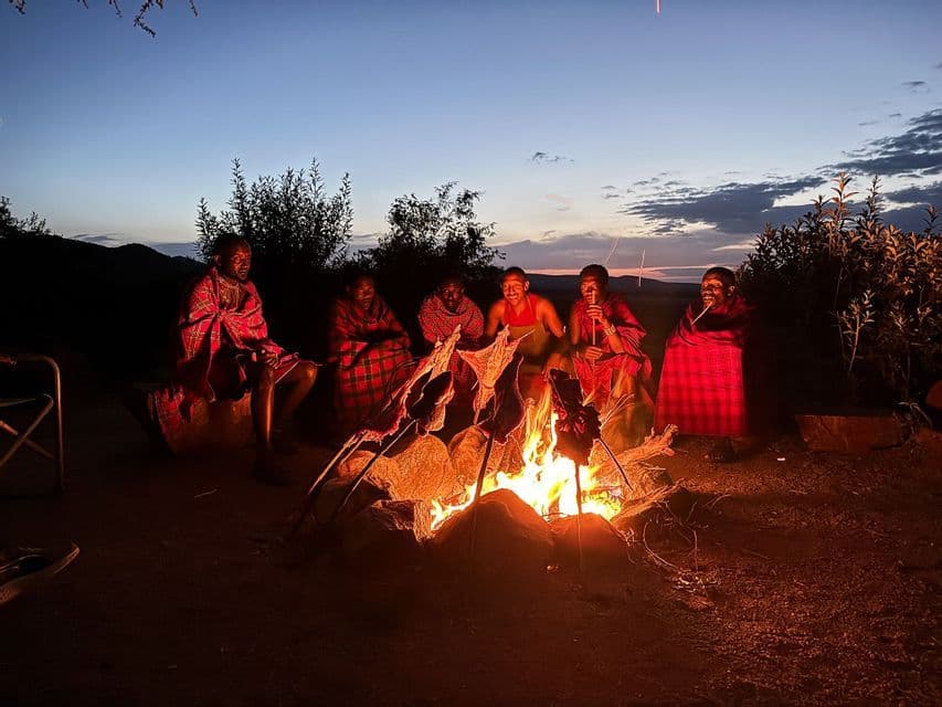 A WeRoad group trip and local people cooking meat skewers over a campfire at dusk.