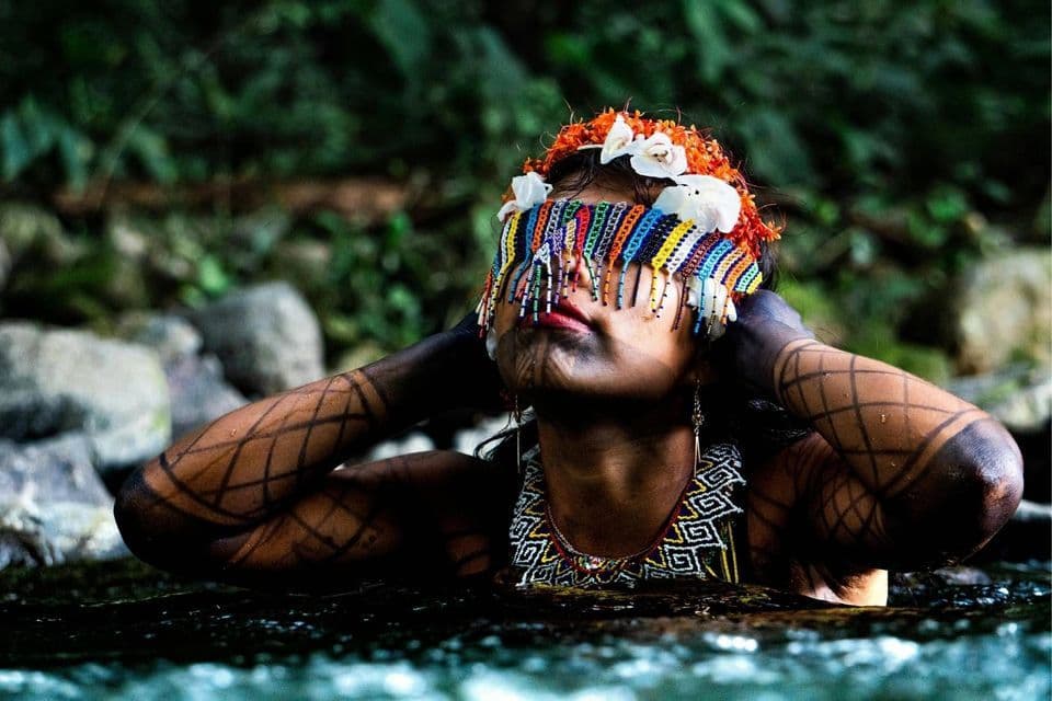 Eine Frau mit geometrischer Körperbemalung und einem farbenprächtigen, perlenbesetzten Kopfschmuck, der ihre Augen bedeckt, badet in einem Fluss in einem üppigen Wald.