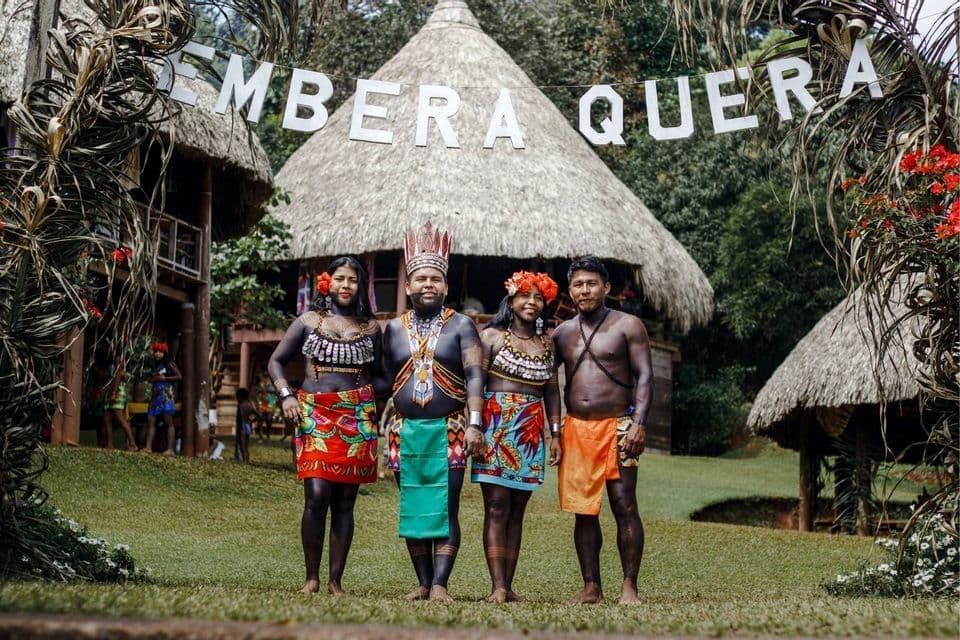 Vier Personen der Embera-Quera-Gemeinschaft in traditioneller Kleidung und Körperbemalung posieren vor Strohhütten.