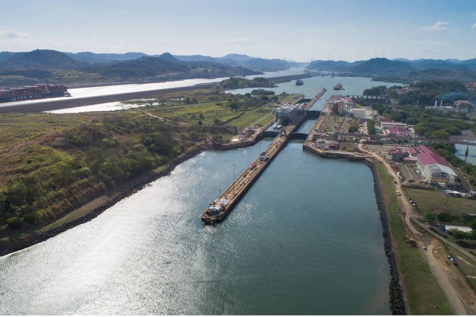 Luftaufnahme eines großen Kanalschleusensystems mit mehreren Frachtschiffen, die die Wasserstraße befahren, umgeben von grünen Hügeln und Infrastruktur.