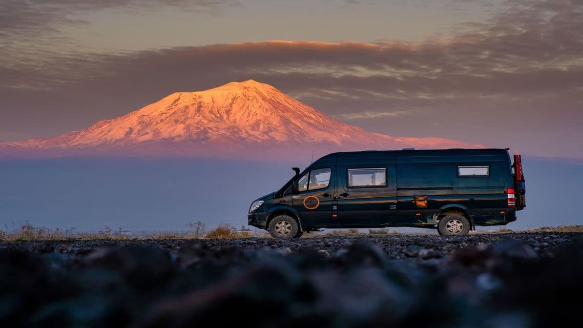 Un camper verde scuro parcheggiato su una strada rocciosa, con una cima montuosa innevata che risplende sullo sfondo all'alba.
