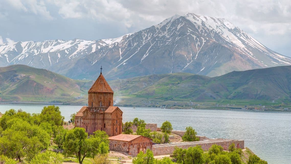 Una storica chiesa in pietra su una verde penisola che si affaccia su un lago, con una montagna innevata sullo sfondo.