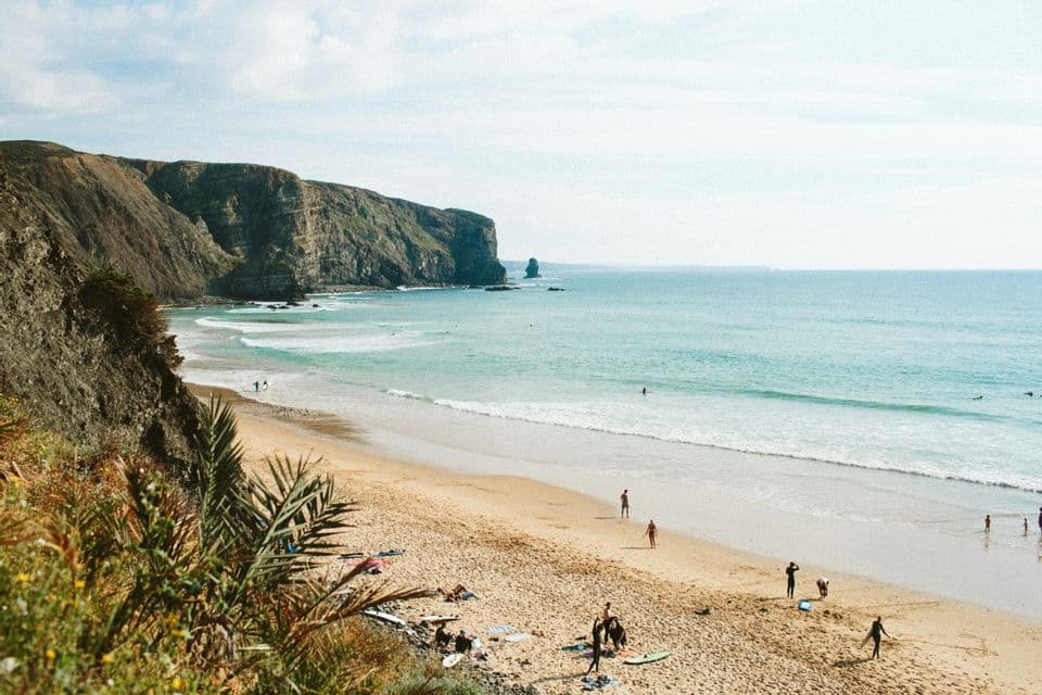 An elevated view of a sandy beach with people relaxing and swimming in the turquoise ocean next to a large, rocky cliff.