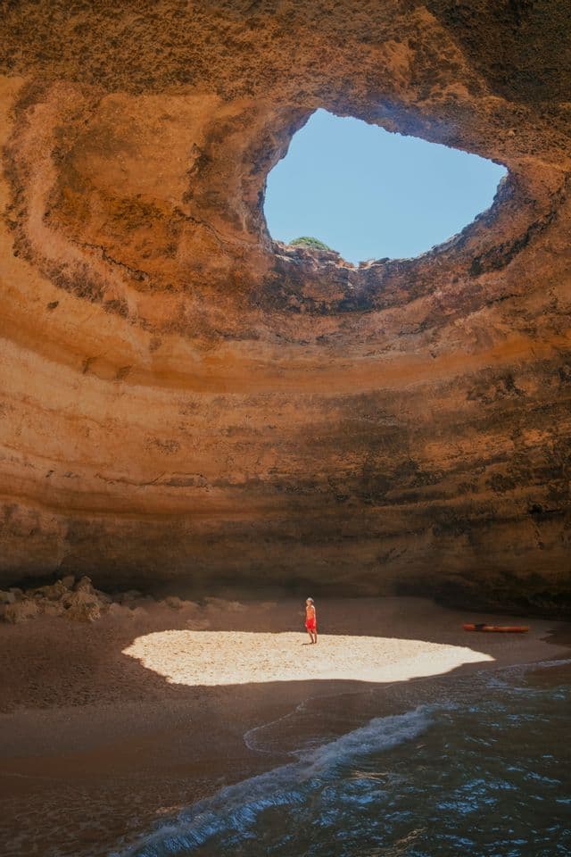 A person in red stands on a patch of sunlit sand inside a large sea cave with an oculus opening to the blue sky.