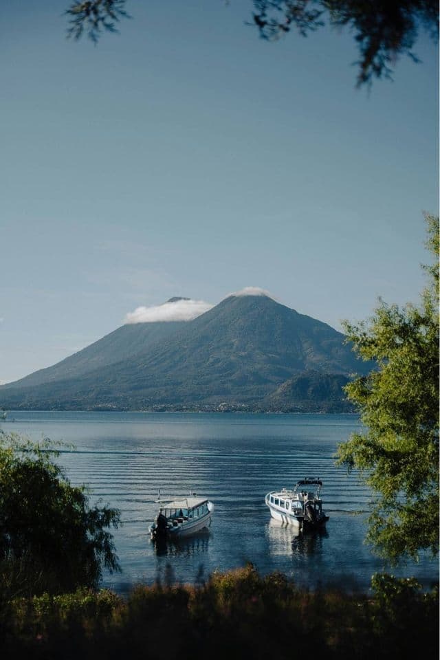Zwei Boote treiben auf einem ruhigen See mit einem großen, wolkenverhangenen Berg im Hintergrund, gesehen durch Bäume.