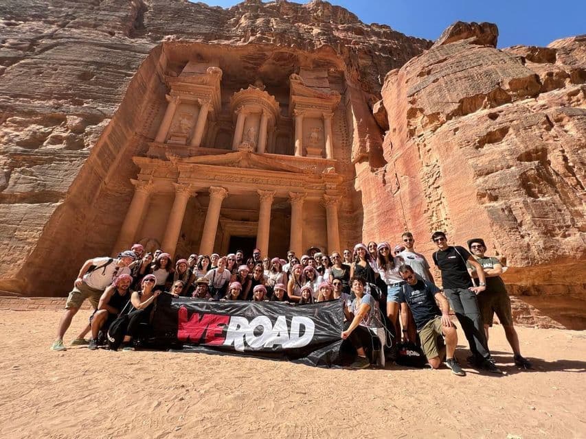 Un grand groupe WeRoad en voyage posant pour une photo avec une bannière devant un temple antique taillé dans une falaise de roche rouge.