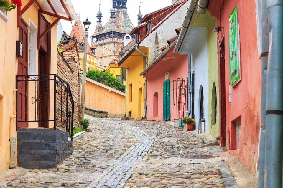 Une rue pavée étroite et déserte, serpentant en montée, bordée de bâtiments colorés, vers un clocher dans une ville historique.