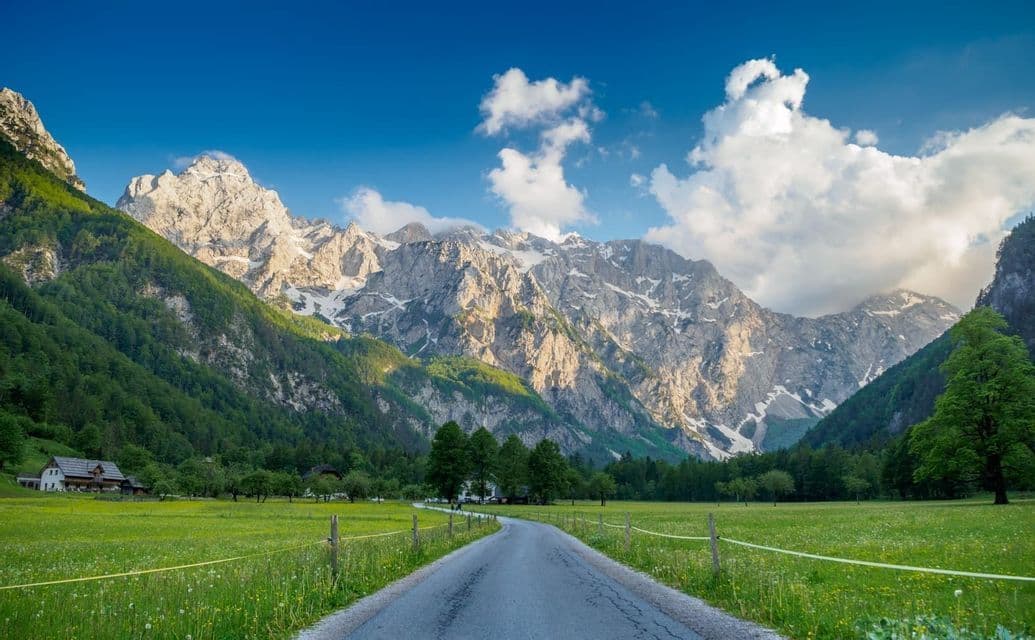 Une route pavée traverse une vallée verdoyante vers une chaîne de montagnes enneigée et ensoleillée, sous un ciel bleu nuageux.