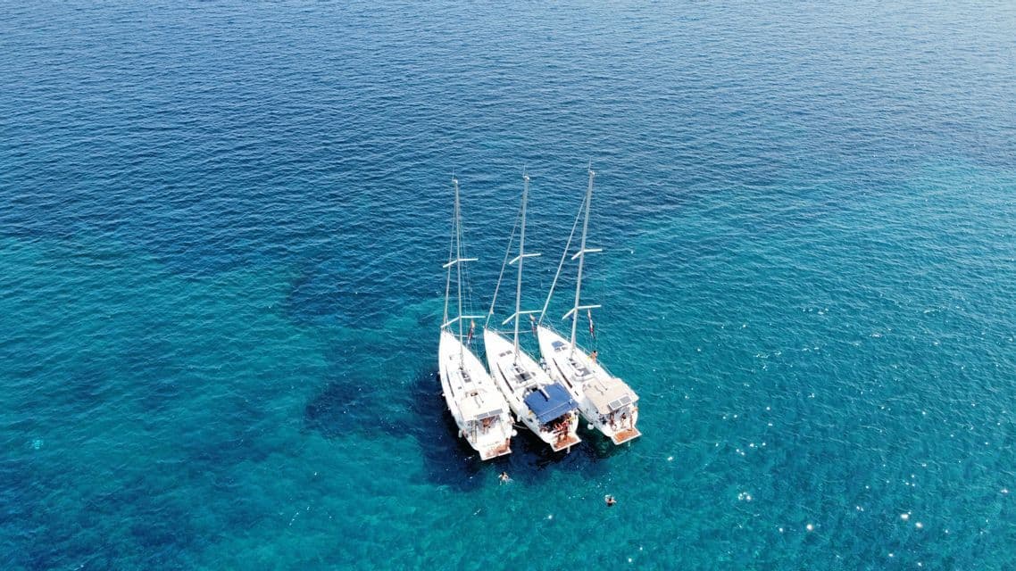 Eine Luftaufnahme von drei Segelbooten einer WeRoad-Gruppenreise, die gemeinsam im klaren türkisfarbenen Wasser ankern, während Menschen in der Nähe schwimmen.