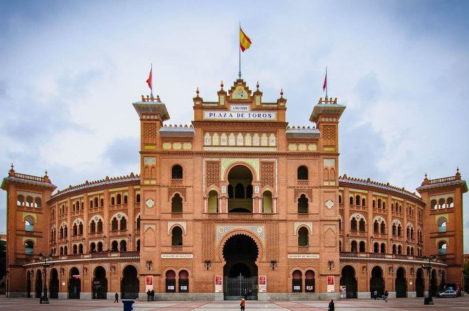 The ornate, reddish-brick facade of the Plaza de Toros bullring, viewed from a large square under a cloudy sky.