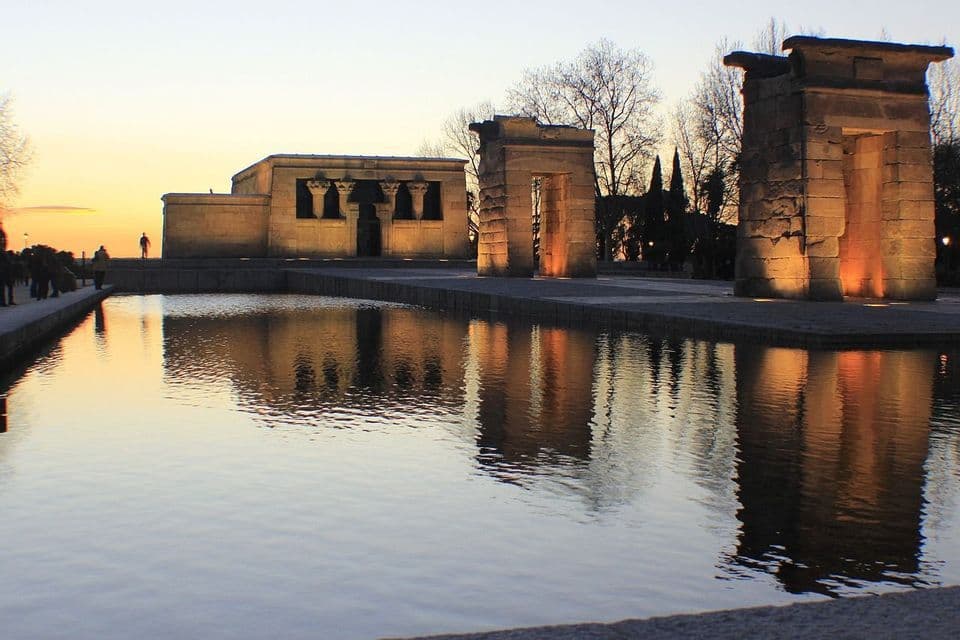 An ancient stone temple and gateways illuminated at sunset, their reflections cast on a calm pool of water.