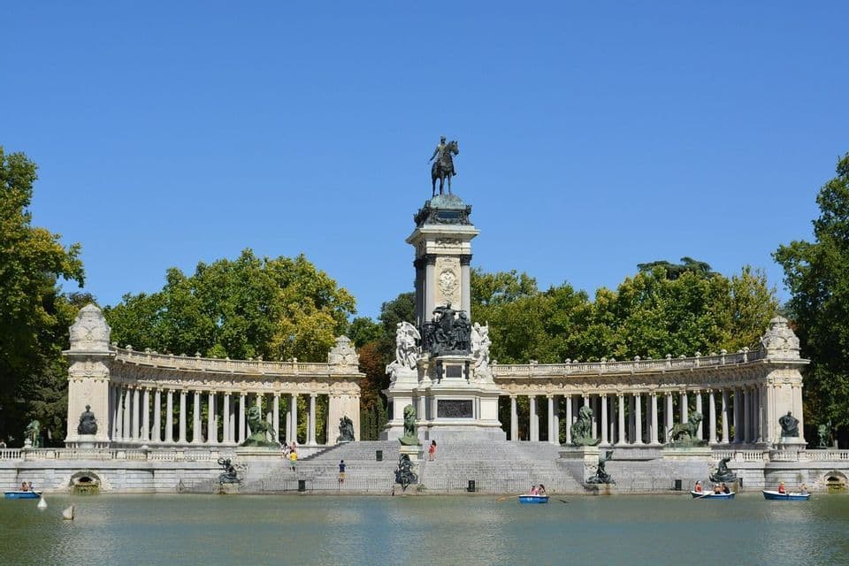 An ornate monument with an equestrian statue and colonnade stands by a lake where people row boats on a sunny day.