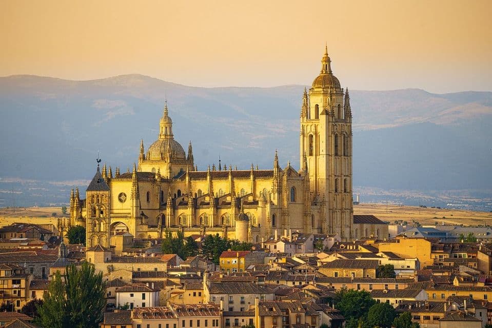A Gothic cathedral bathed in golden sunset light, overlooking a town with distant mountains in the background.