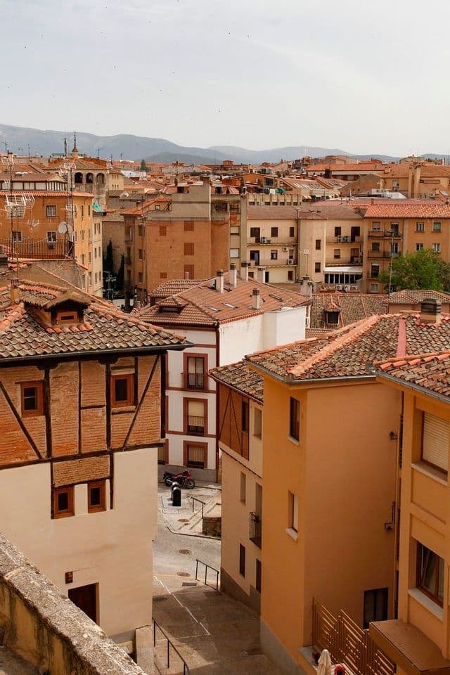 A high-angle view of a town with terracotta-tiled roofs, with a narrow street below and mountains in the background.