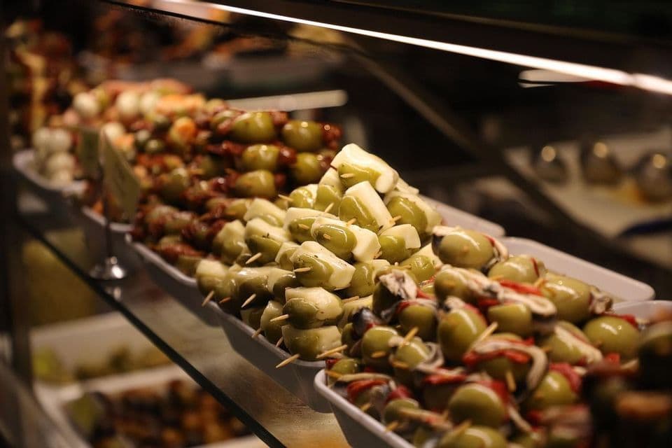 A close-up of various tapas on skewers, featuring green olives with cheese and peppers, arranged in a food display case.