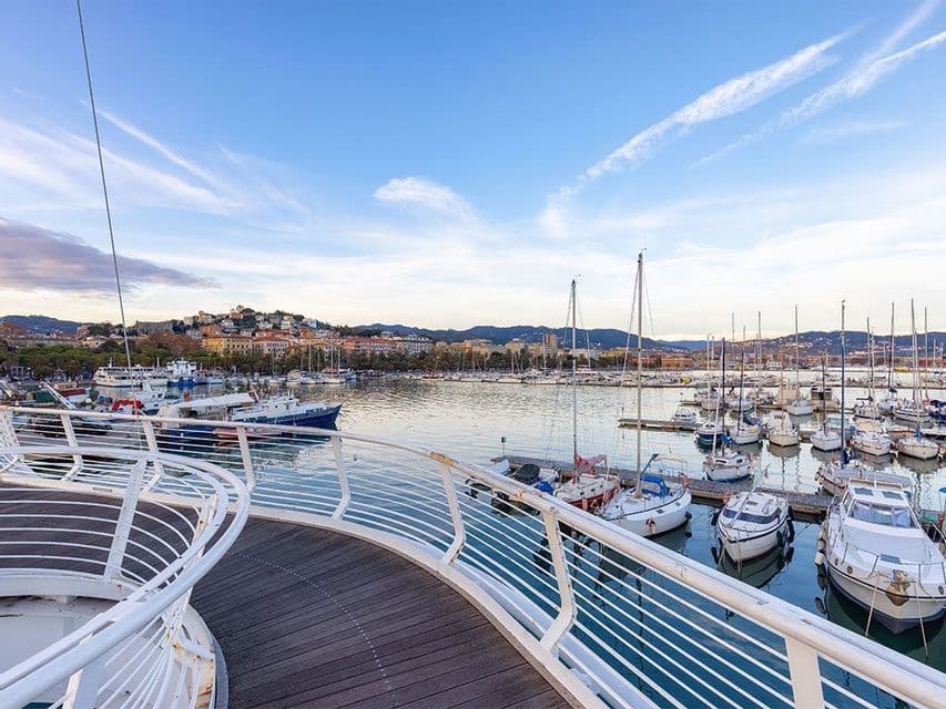 Una passerella curva in legno con ringhiere bianche si affaccia su un porto pieno di barche a vela, con una cittadina collinare sullo sfondo.