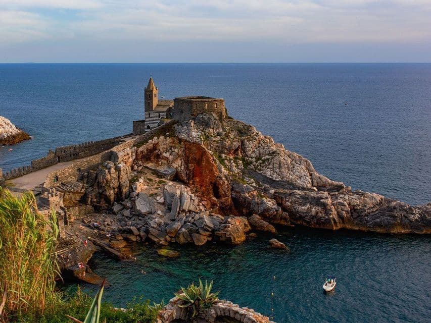 Una chiesa in pietra con un campanile sorge su un promontorio roccioso che domina il mare blu, con una piccola barca che galleggia in basso.