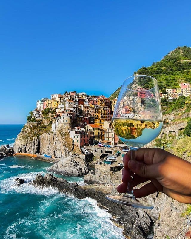 Una mano sostiene una copa de vino blanco frente a un pueblo colorido construido en un acantilado con vistas a un mar turquesa.