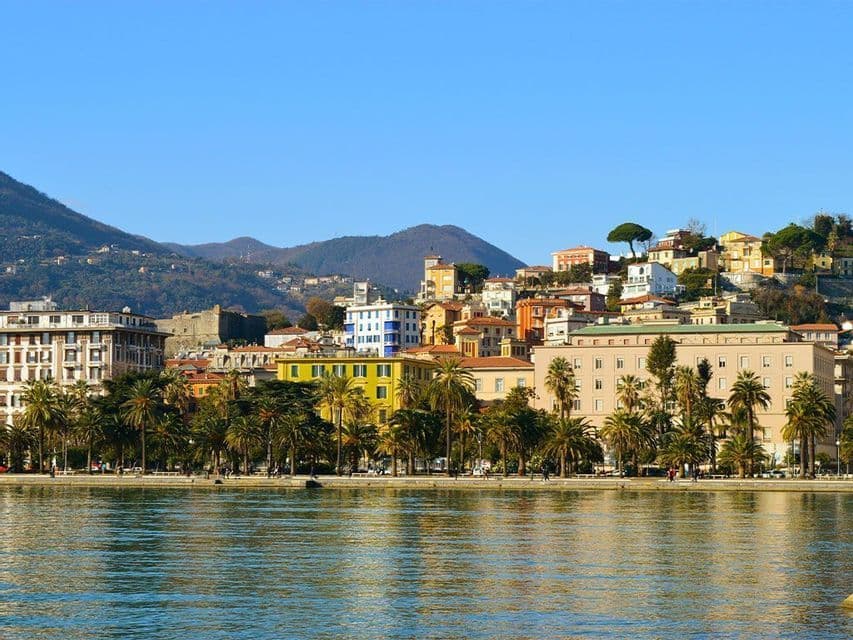 Une vue sur le front de mer d'une ville côtière avec des bâtiments colorés et des palmiers, adossée à des collines verdoyantes sous un ciel bleu clair.