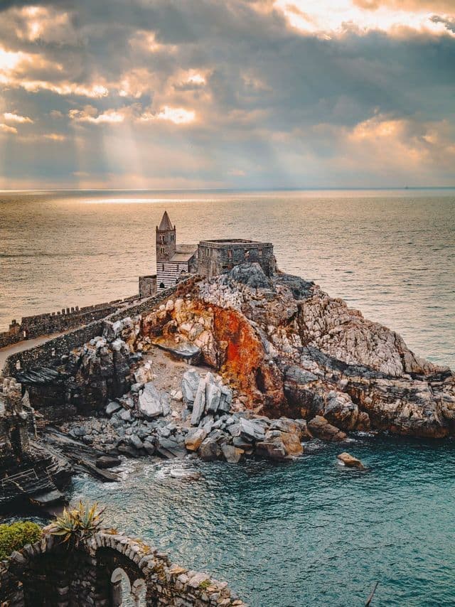 Una chiesa di pietra su una scogliera rocciosa a picco sul mare, con raggi di sole che filtrano tra le nuvole sovrastanti.