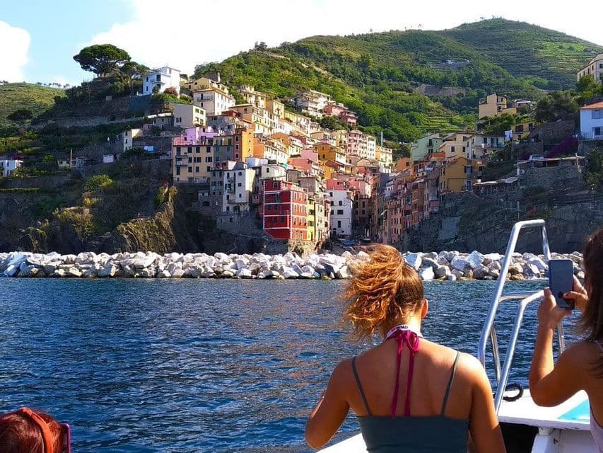 Dos mujeres de un viaje en grupo de WeRoad en un barco, observando un colorido pueblo en acantilado, mientras una de ellas toma una foto.