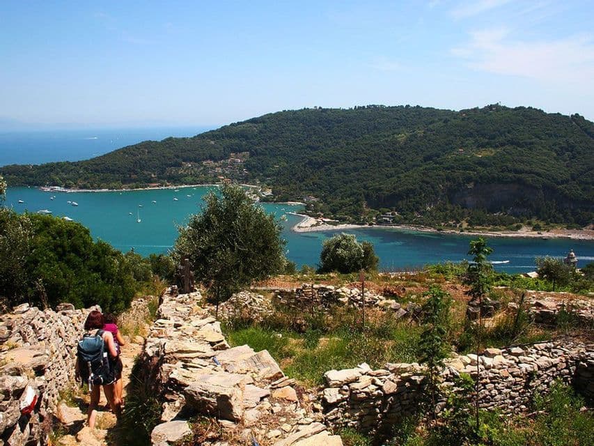 Zwei Wanderer mit Rucksäcken gehen einen felsigen Küstenpfad entlang, mit Blick auf eine türkisfarbene Bucht und eine bewaldete Halbinsel.