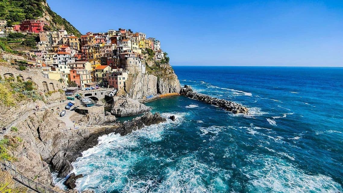 Case colorate su una scogliera si affacciano su una baia rocciosa con un mare blu intenso sotto un cielo sereno.