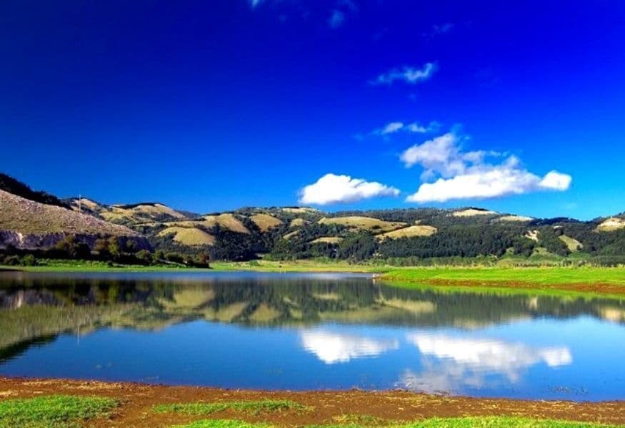 Un lago calmo riflette colline ondulate ed erbose e un cielo blu intenso con nuvole bianche in una giornata di sole.