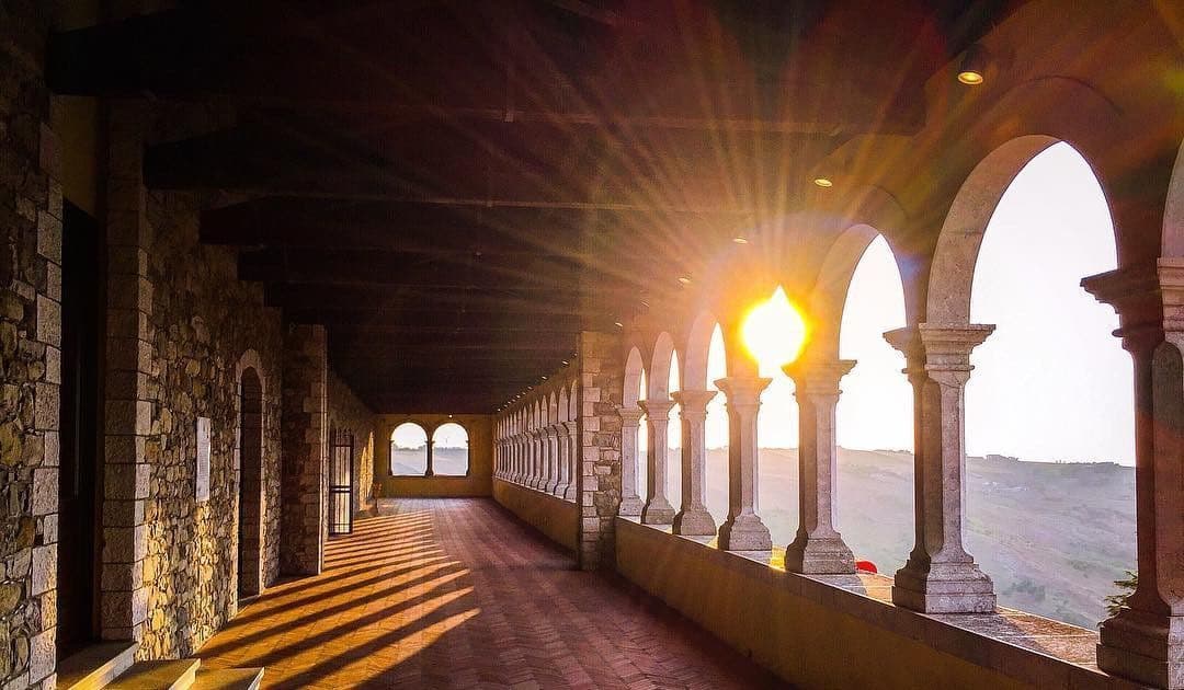 Un lungo colonnato in pietra con archi che si affaccia su un paesaggio collinare al tramonto, proiettando lunghe ombre sul pavimento piastrellato.