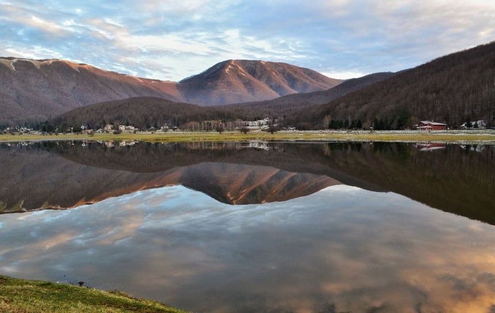 Montagne e un cielo nuvoloso si specchiano perfettamente in un lago calmo, con un piccolo villaggio visibile sulla sponda opposta.