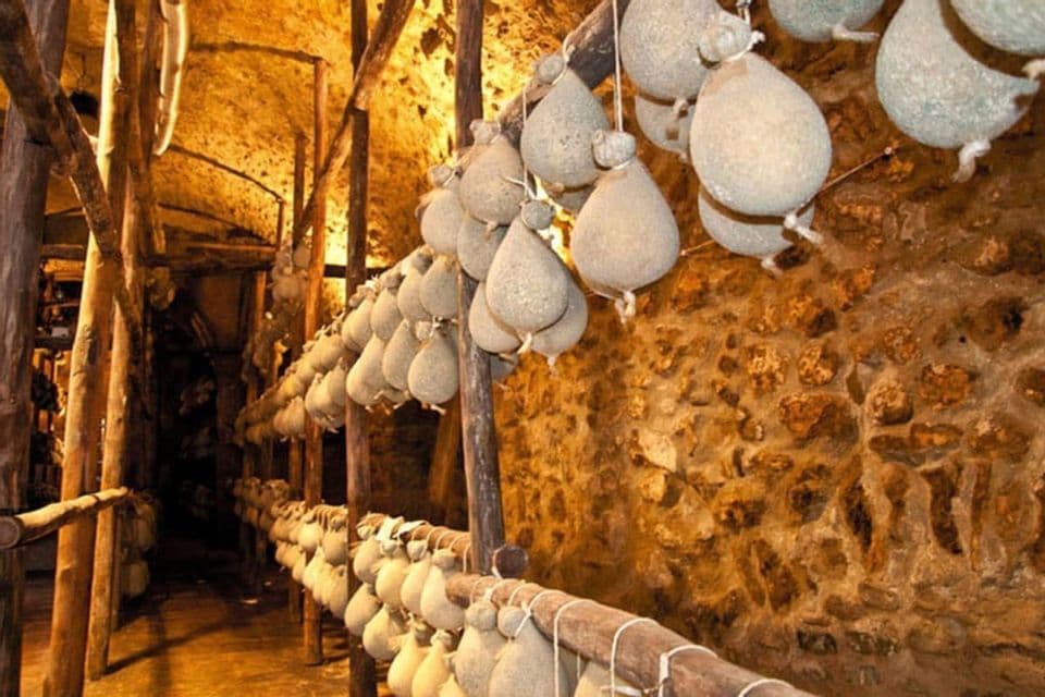Fila di formaggi a forma di lacrima appesi a rastrelliere di legno per stagionare all'interno di una rustica cantina in pietra.