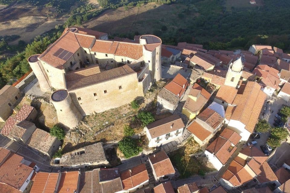 Una vista aerea di un castello di pietra con torri circolari, immerso in un borgo collinare con tetti in tegole di terracotta sotto il sole.