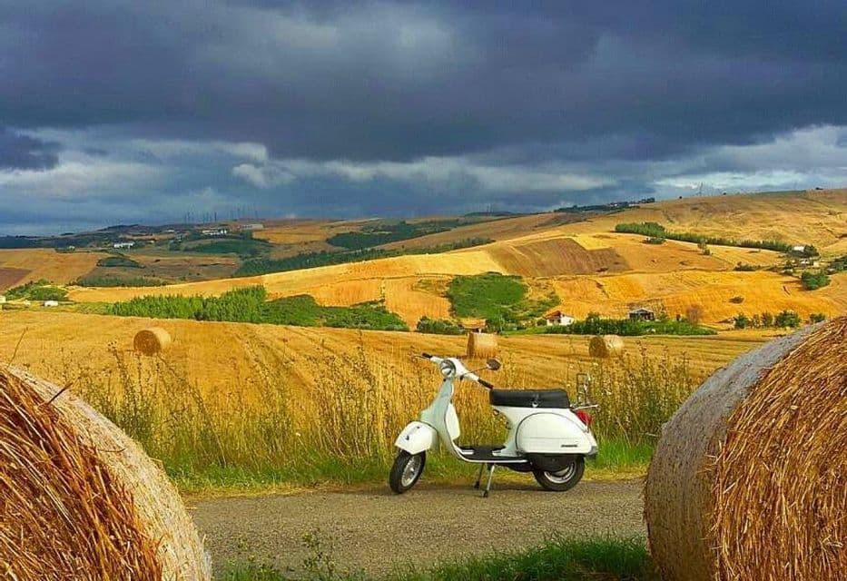 Uno scooter bianco parcheggiato su una strada tra balle di fieno in un paesaggio collinare con campi mietuti sotto un cielo tempestoso.