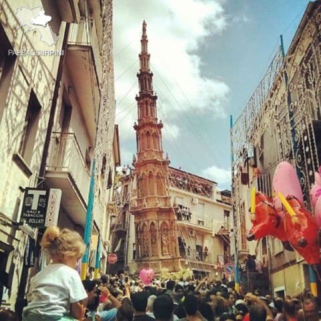 Una grande folla ammira un alto e sontuoso obelisco processionale che si muove lungo una stretta via decorata durante una festa.