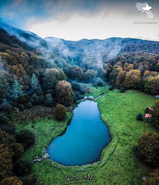 Una vista aerea di un piccolo lago blu in un prato verde, circondato da una fitta foresta autunnale con nebbia che si estende sulle colline lontane.